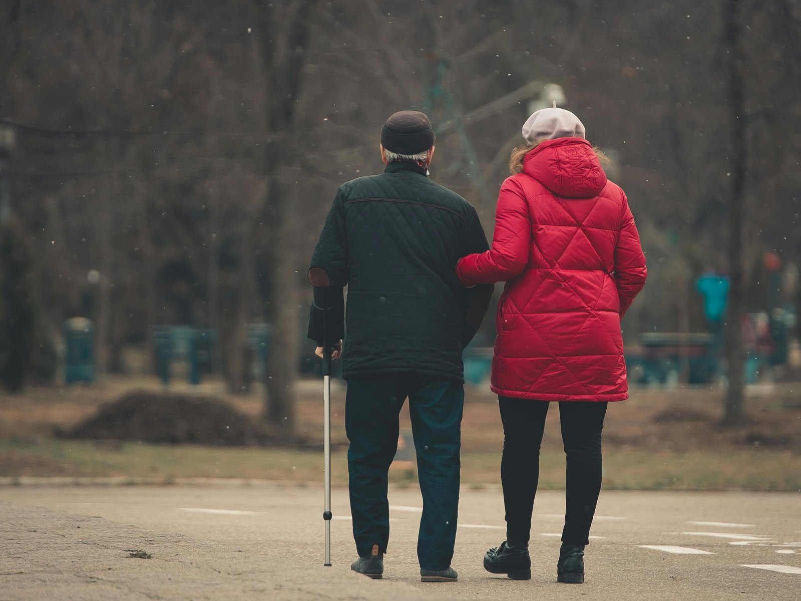 An elderly couple walks arm in arm with a walking stick in a park.